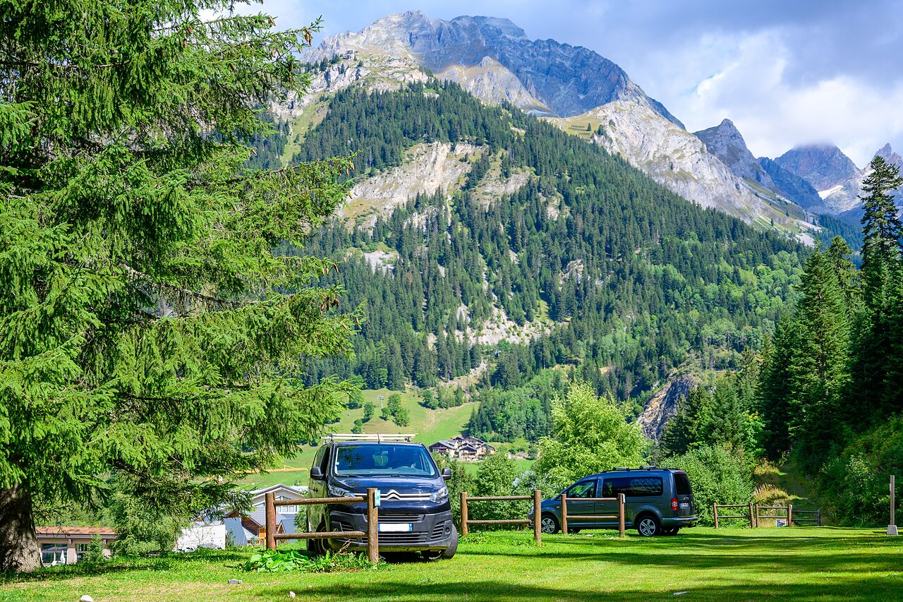 Vans parked on grassy pitch with mountains and forest at CLICOCHIC Alpes Lodges campsite in Pralognan la Vanoise (73).