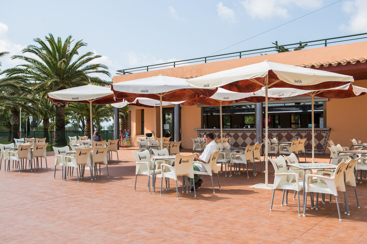 Outdoor restaurant-bar terrace with tables, umbrellas, and palm trees at CAPFUN Aube campsite in Deltebre, Tarragona (43).