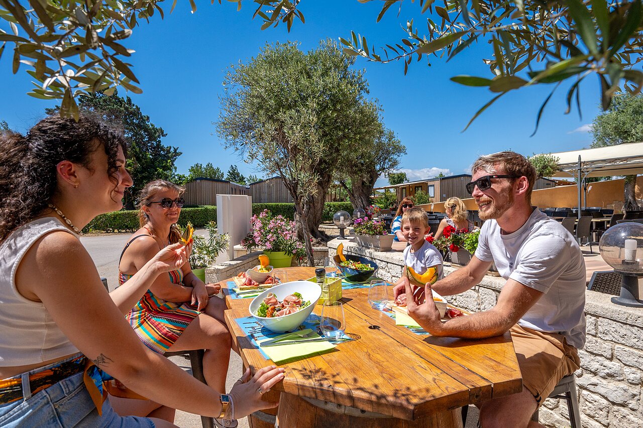 Smiling family dining on shaded terrace, mobile homes at CAPFUN Barbacane campsite in NARBONNE (11).