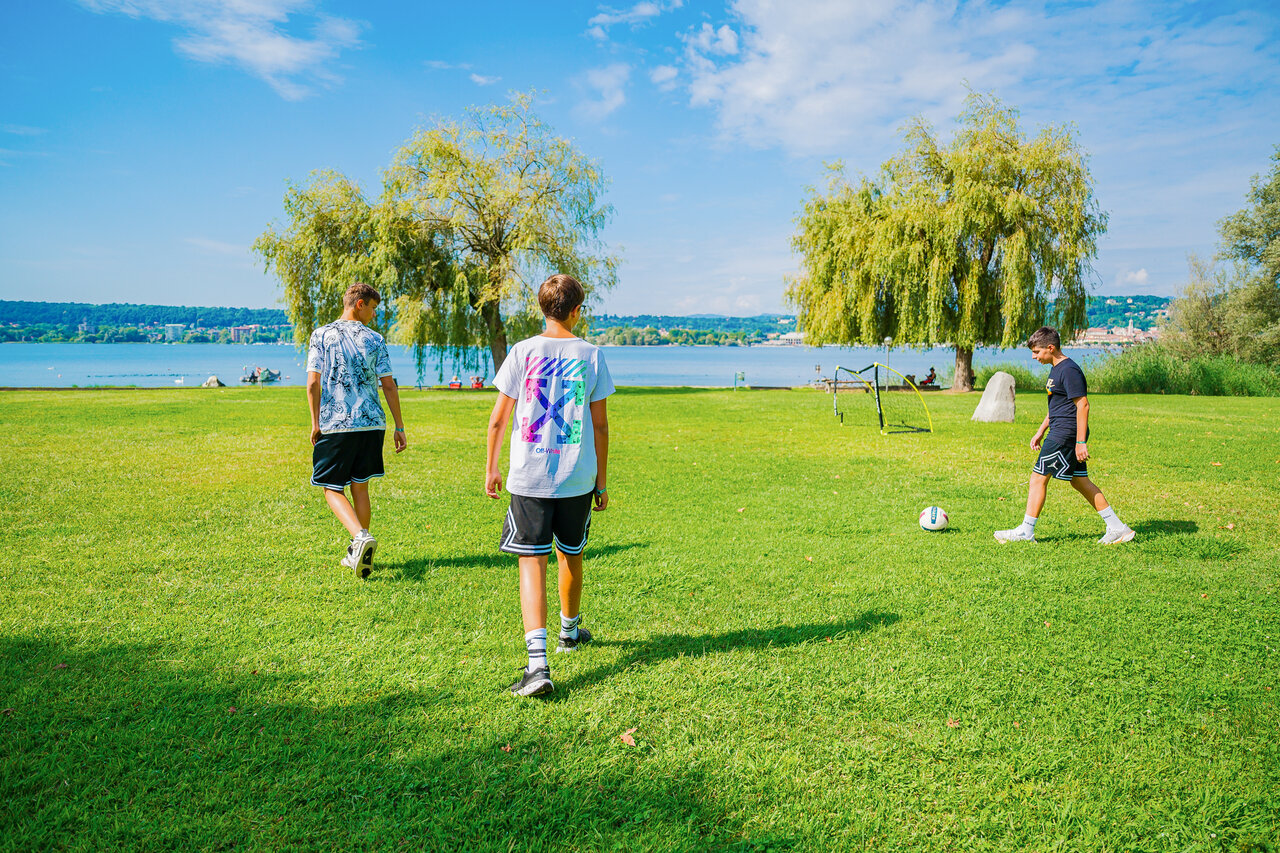 Boys playing football on grass, lake, CAPFUN Citta di Angera in Angera VA.