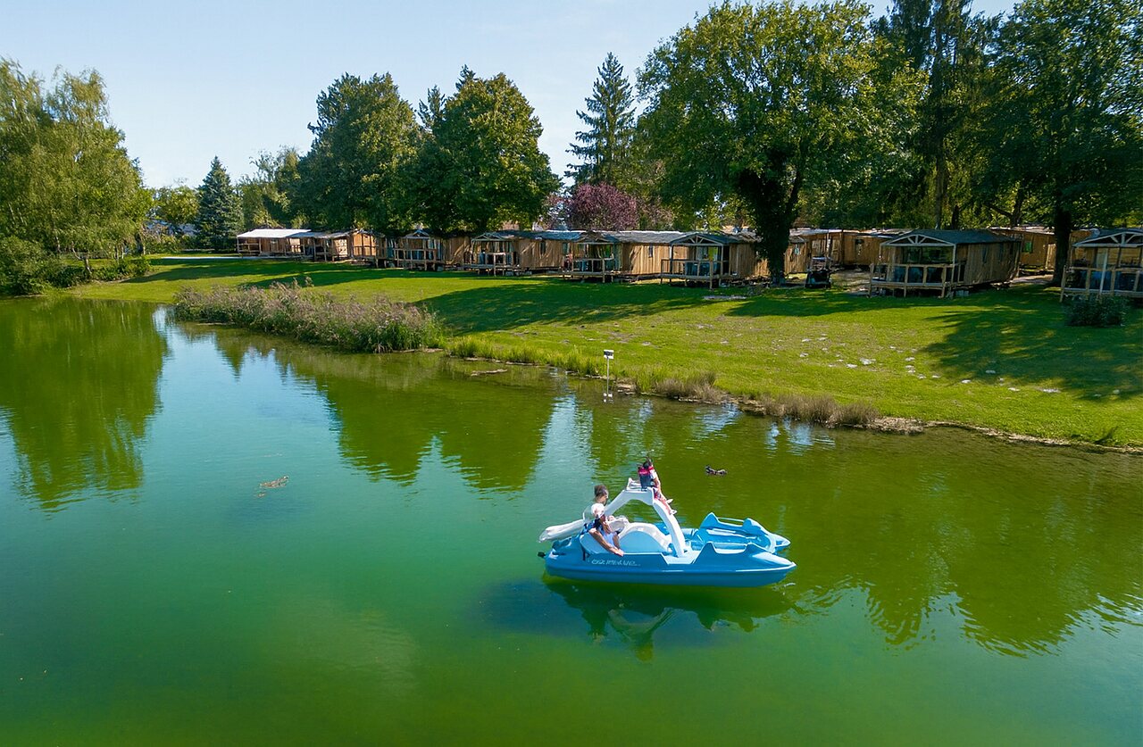 Pedal boat on the lake with wooden accommodations at CLICOCHIC Au Clair Ruisseau campsite in Gerstheim (67).