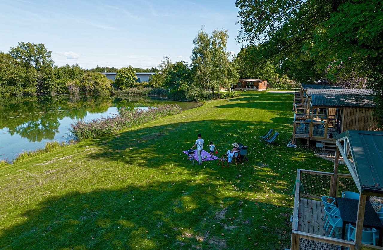 Wooden cabins, lake, and family picnicking at CLICOCHIC Au Clair Ruisseau campsite.