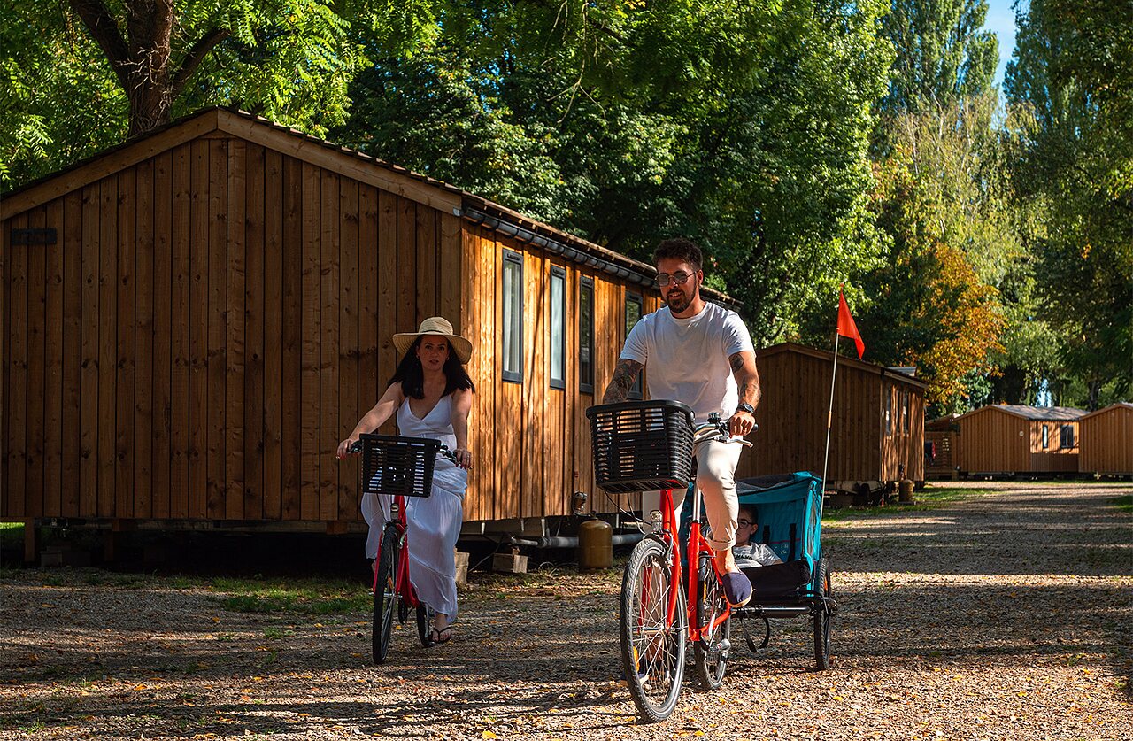 Couple cycling with child trailer in front of accommodations at camping CLICOCHIC Au Clair Ruisseau in Gerstheim (67).