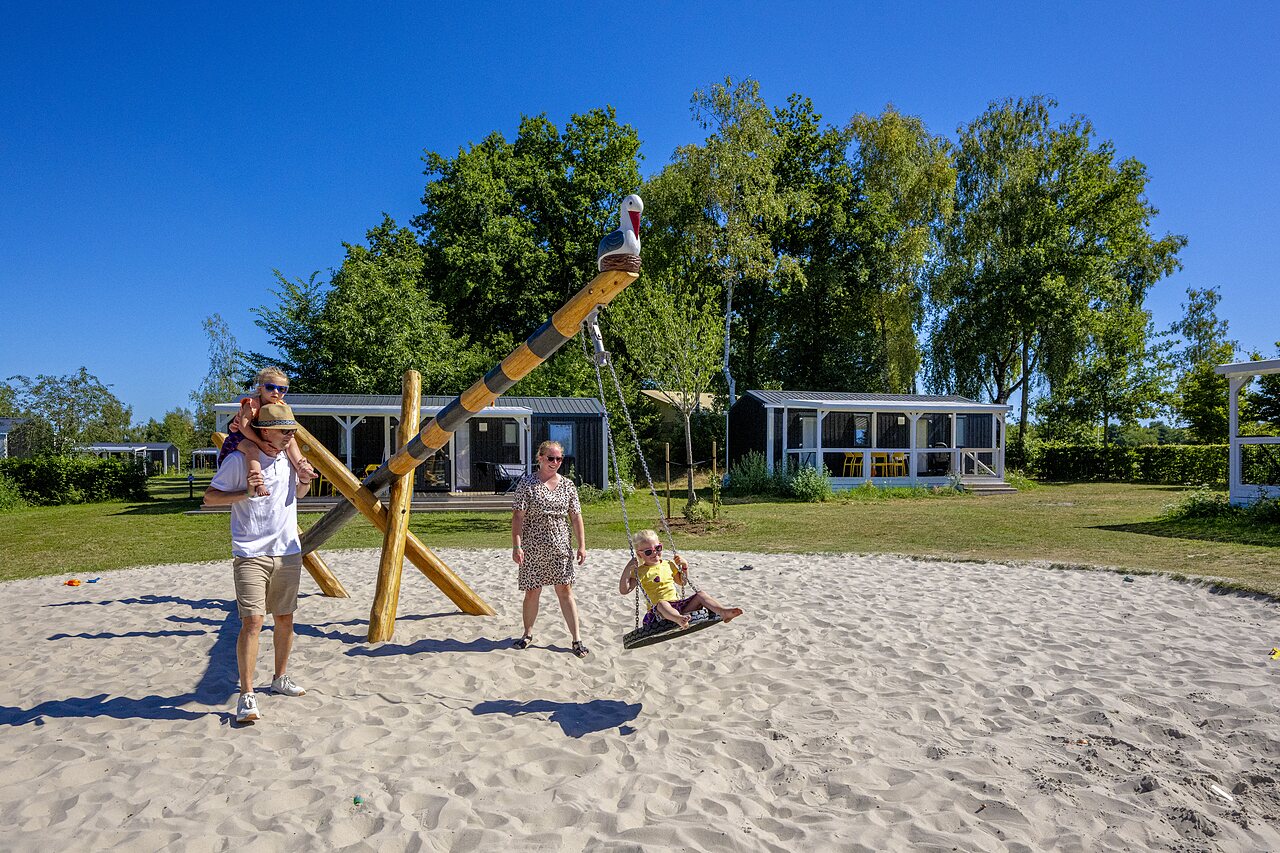 Sandy playground with swing and family, mobile homes at CAPFUN De Belten campsite in Rheeze.