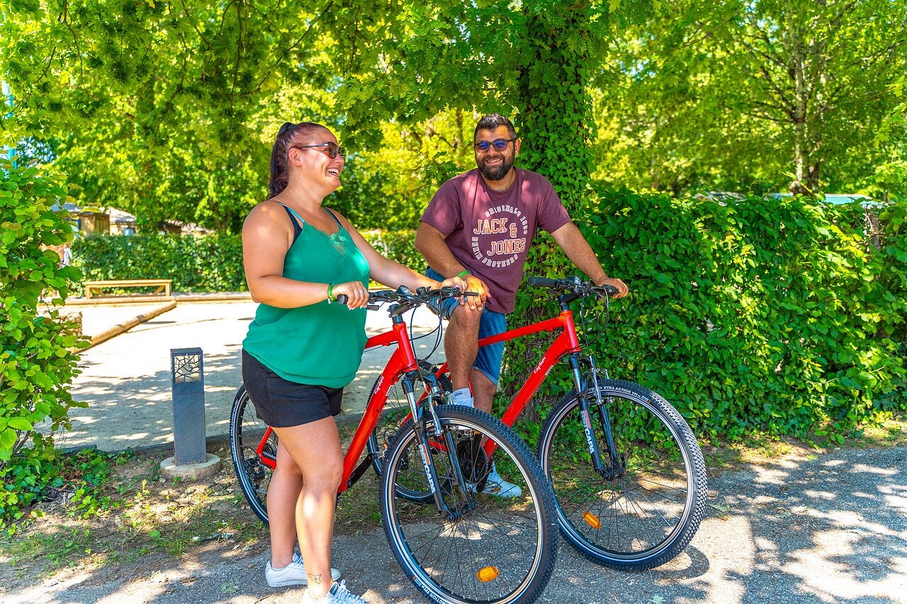 Smiling couple with red bikes, surrounded by greenery, at CAPFUN Duravel campsite in Duravel (46).