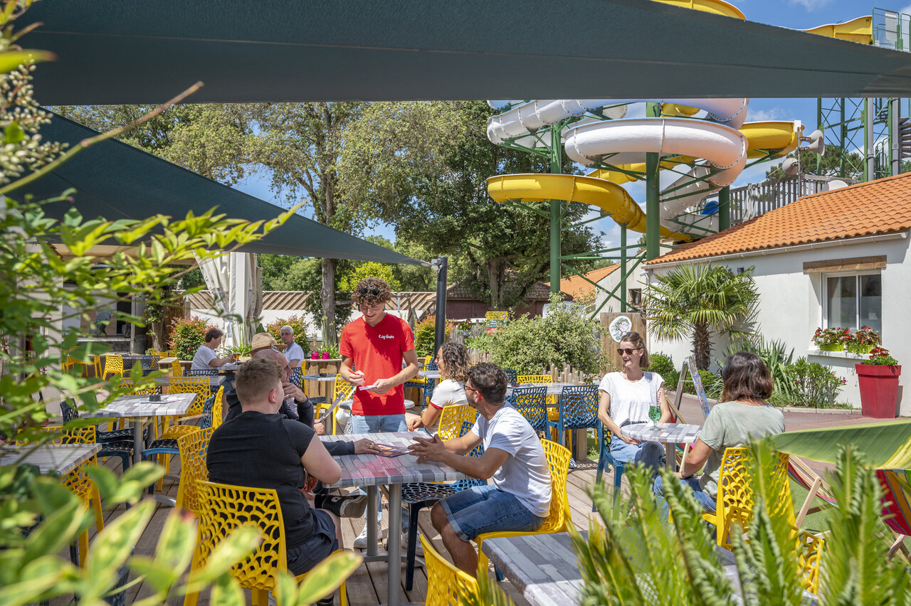 Lively restaurant terrace with water slides in background at CAPFUN Ecureuils campsite in JARD SUR MER (85).