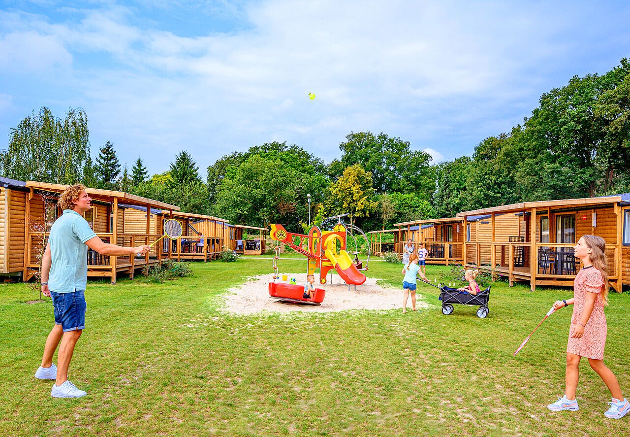 Family playing badminton and children at playground, wooden accommodations at CAPFUN Eibernest campsite in Eibergen.