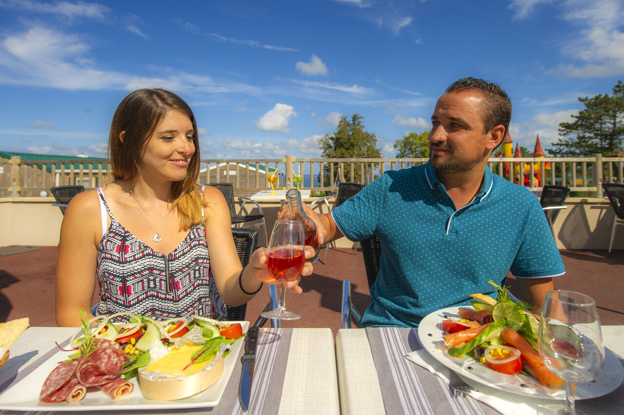 Couple dining on restaurant terrace, toasting ros� wine, at CAPFUN Falaises campsite.