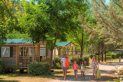 Wooden mobile homes and family walking on a path at CLICOCHIC Filament campsite in Thoiras (30).