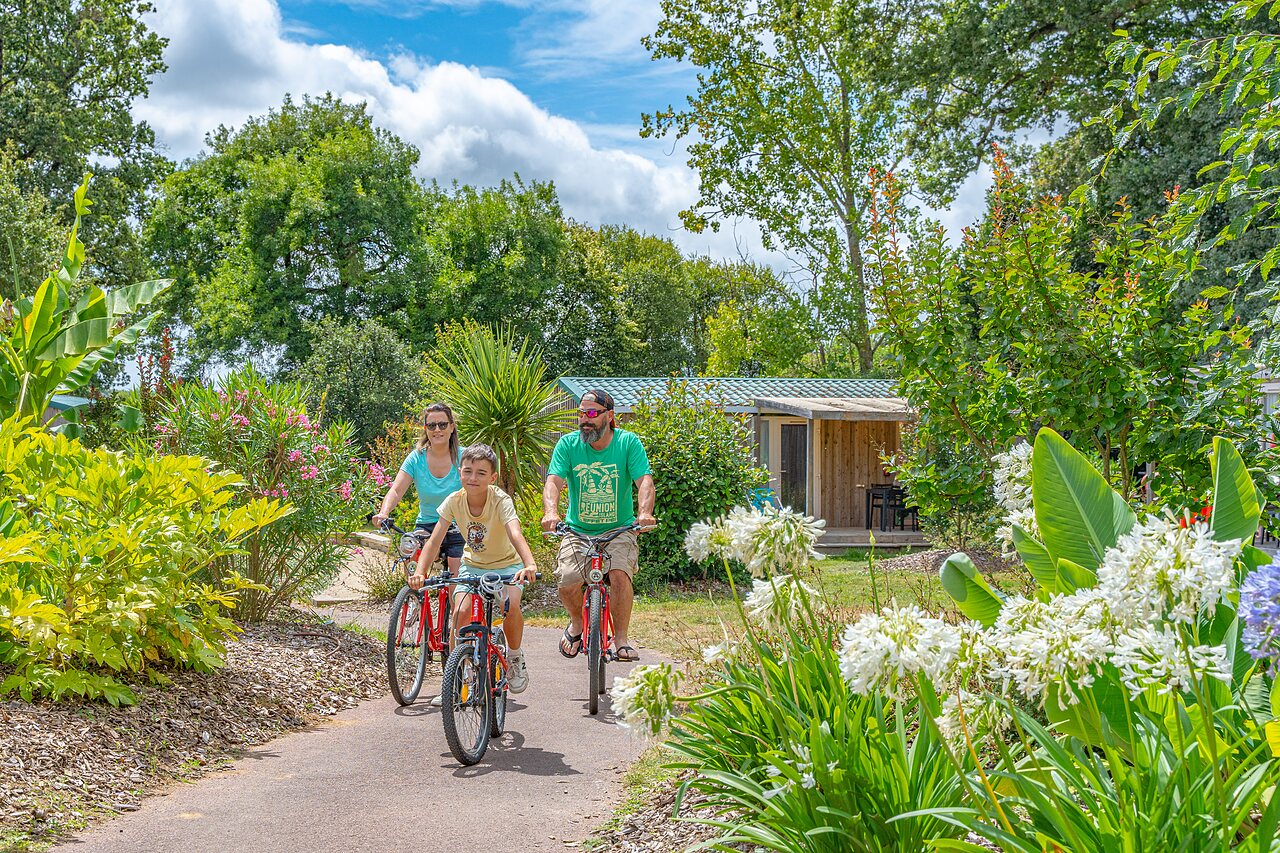 Family cycling on flowery path, accommodation in background at CAPFUN Forges campsite in Avrille (85).