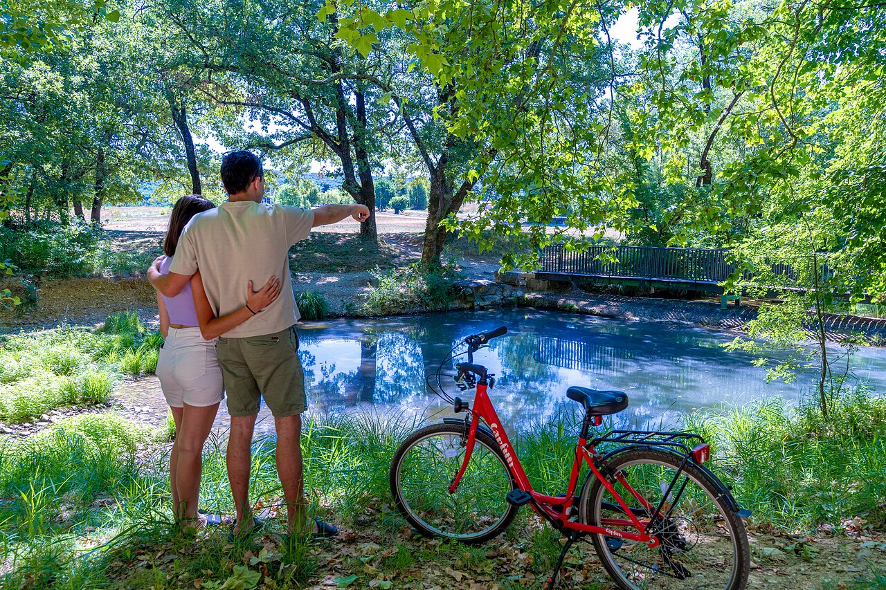 Couple and red bicycle by a wooded pond at CAPFUN Fumades campsite in ALLEGRE-LES-FUMADES (30).