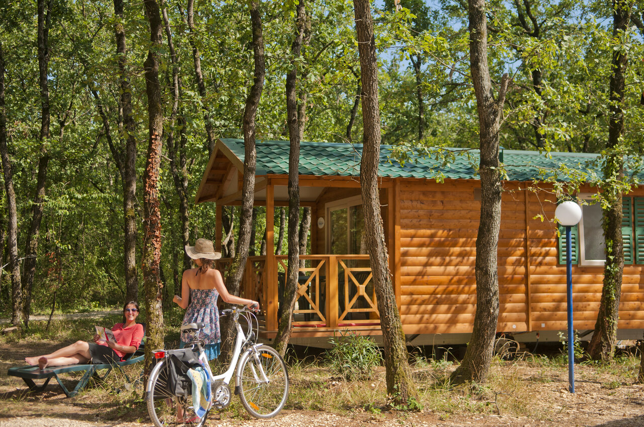 Wooden chalet, woman with bike and another reading, under trees at CAPFUN Grand Lierne campsite in CHATEAUDOUBLE (26).