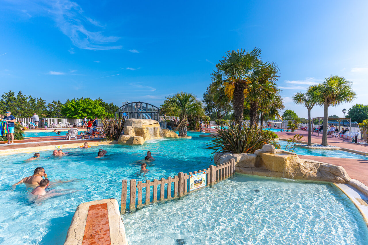 Outdoor swimming pool with waterfall, palm trees and swimmers at CAPFUN Hautes Coutures campsite in BENOUVILLE (14).