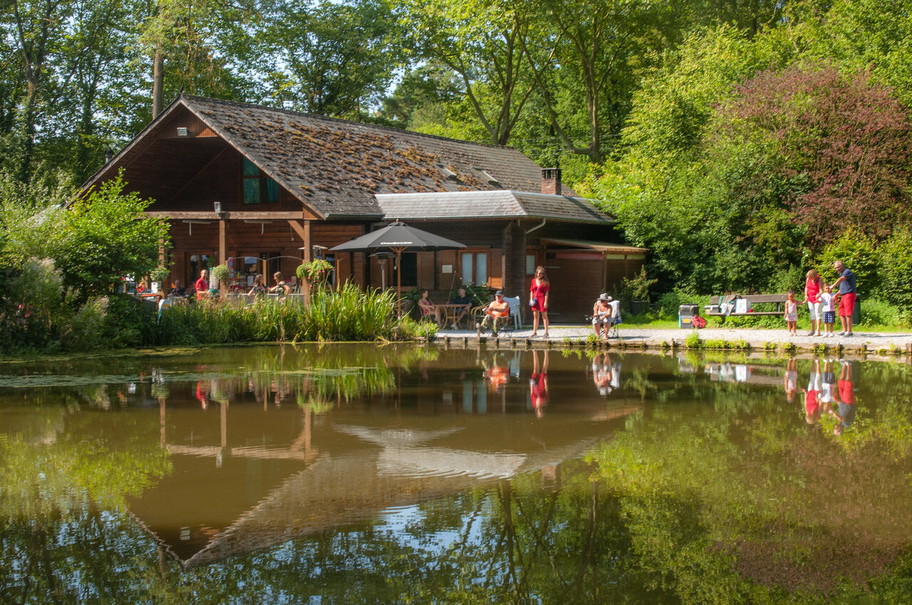 Restaurant building with terrace by the pond, people fishing at CAPFUN Hirondelle campsite in Oteppe.
