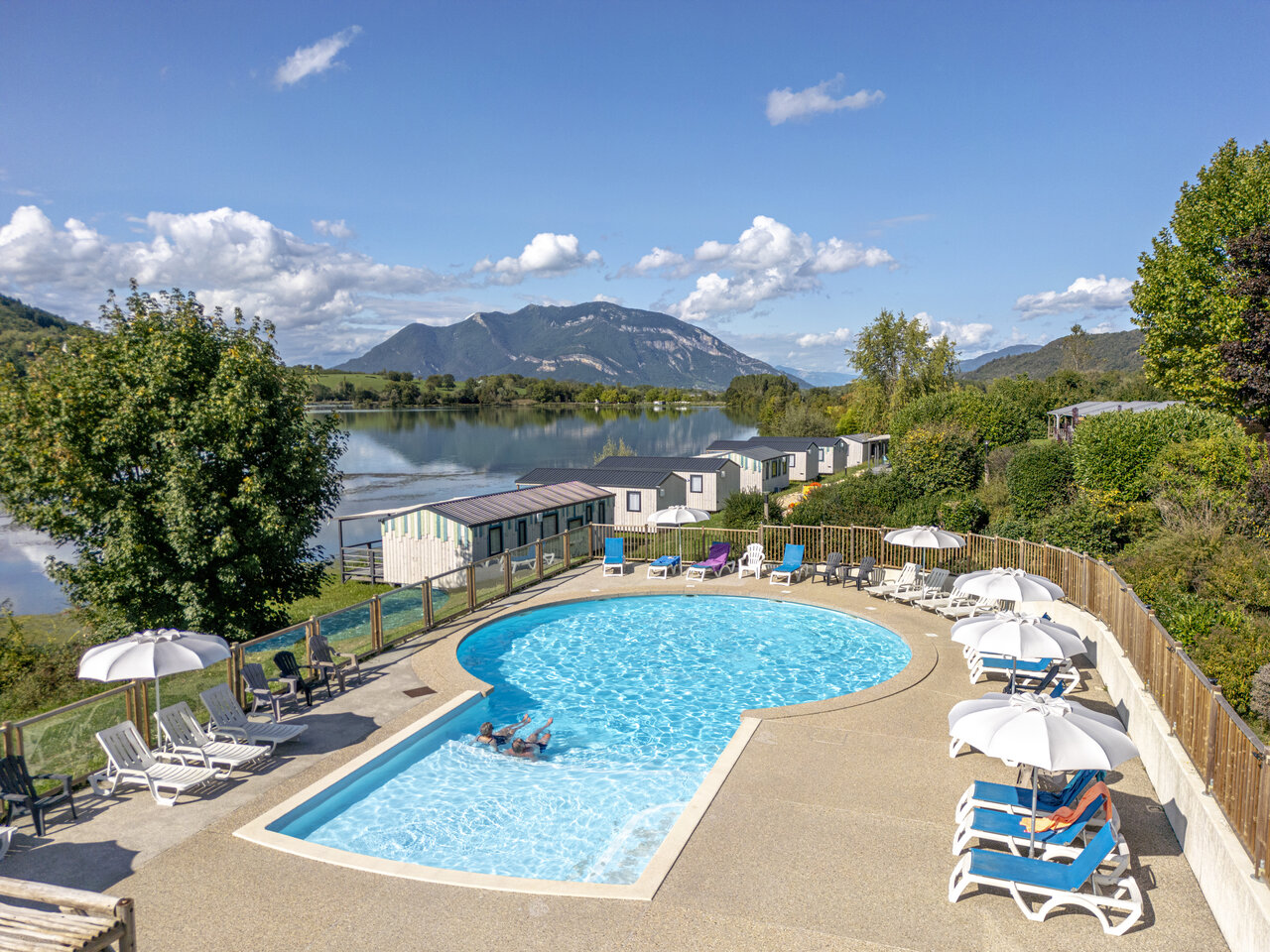 Outdoor swimming pool, sun loungers, lake and mobile homes at CLICOCHIC Lac du Lit du Roi campsite in Massignieu-de-Rives (01).