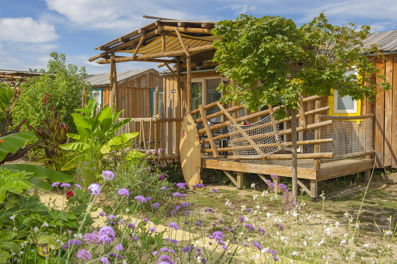 Wooden chalet with raised terrace, surfboard and lush vegetation at CAPFUN Lila campsite in LINXE (40).