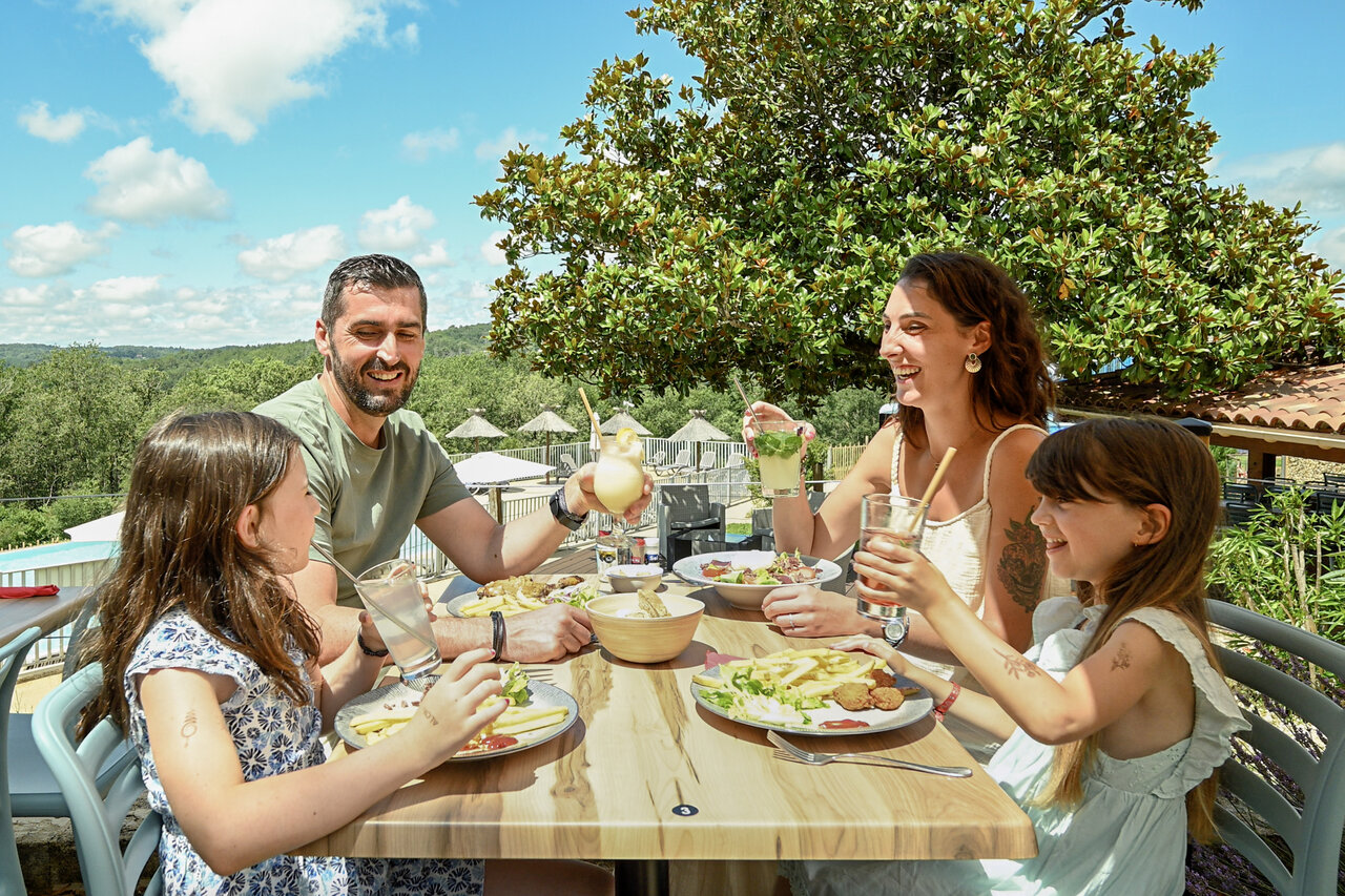 Smiling family enjoying lunch on the restaurant-bar terrace at camping CLICOCHIC Linotte in Le Bugue.
