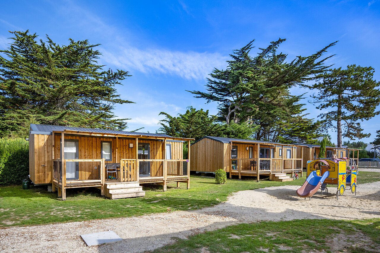 Wooden mobile homes, terraces and children's playground at CLICOCHIC Longchamp campsite.