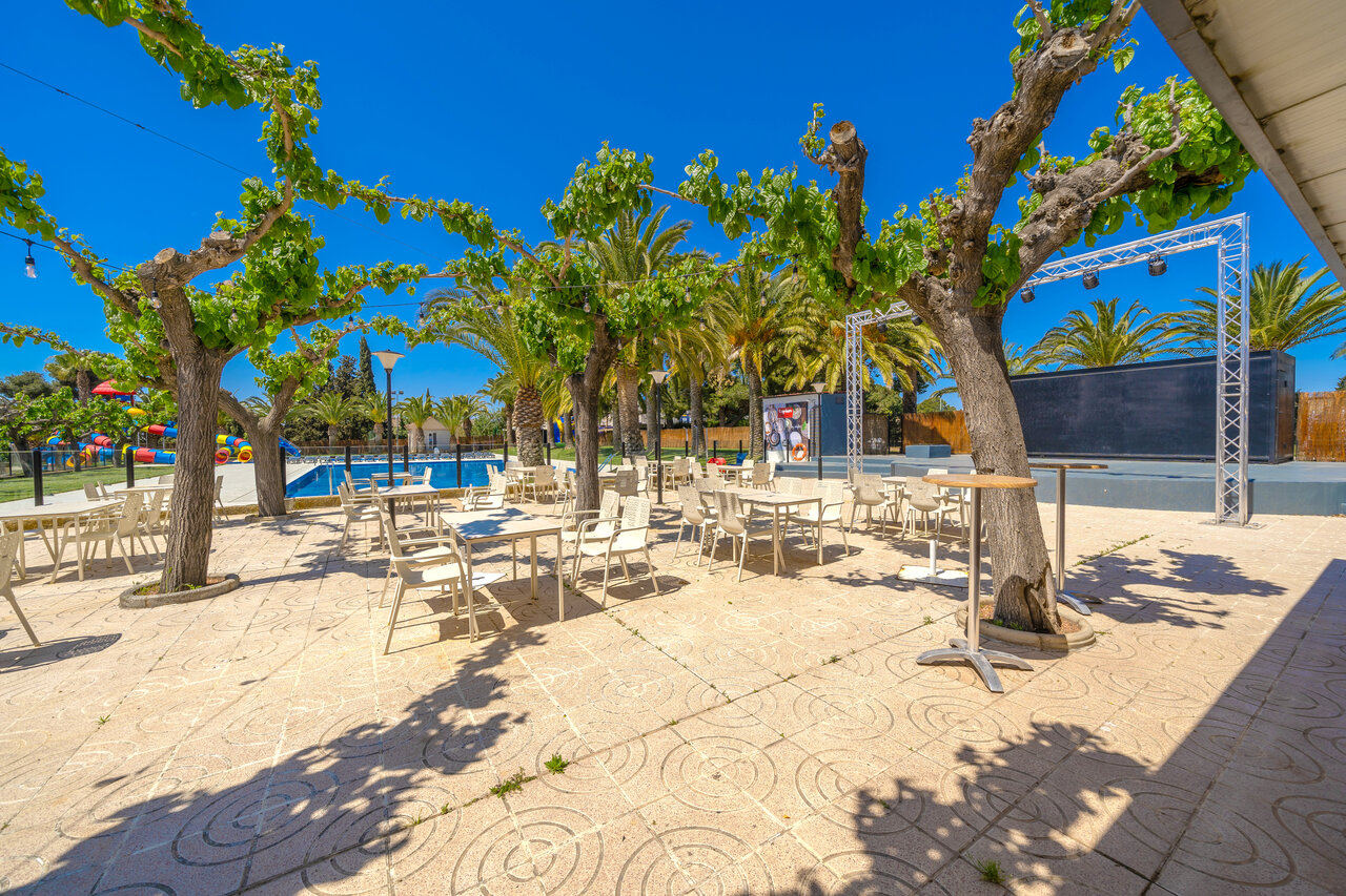 Shaded restaurant terrace, swimming pool and stage at CAPFUN Mirmanda campsite in Cambrils.