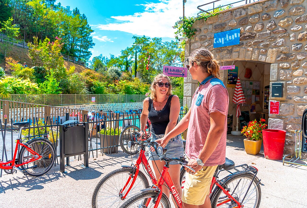 Two smiling people with red bicycles near the swimming pool at CAPFUN Plantas campsite in SAINT VINCENT DE DURFORT (07).