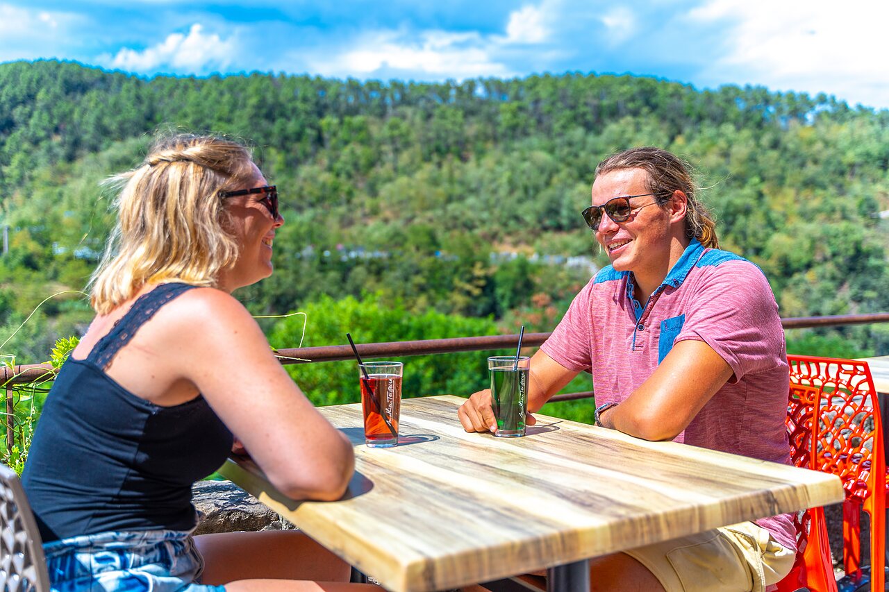 Outdoor bar, couple with drinks, at CAPFUN Plantas campsite in SAINT VINCENT DE DURFORT (07).