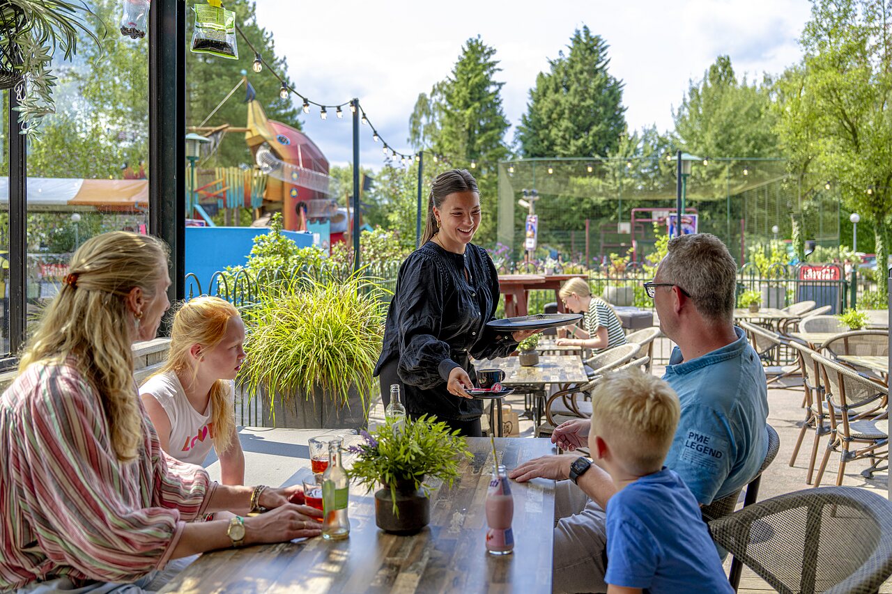 Family dining on terrace, waitress, children's playground at CAPFUN Ponderosa campsite in Ulicoten.