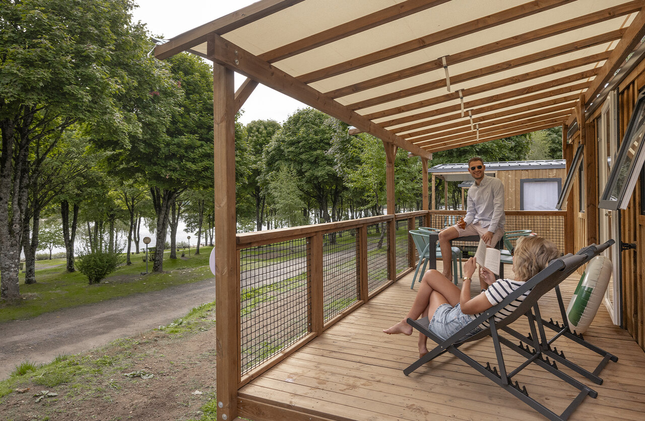 Couple on wooden Mobil-home terrace, lake view, at CLICOCHIC Soleil Levant campsite.