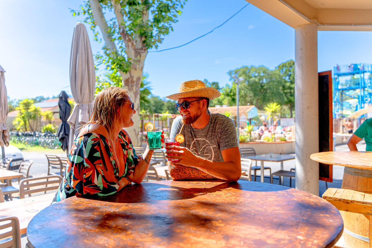 Smiling couple drinking cocktails at the bar, pool in background, at CLICOCHIC Texas campsite in Argel�s sur Mer (66).