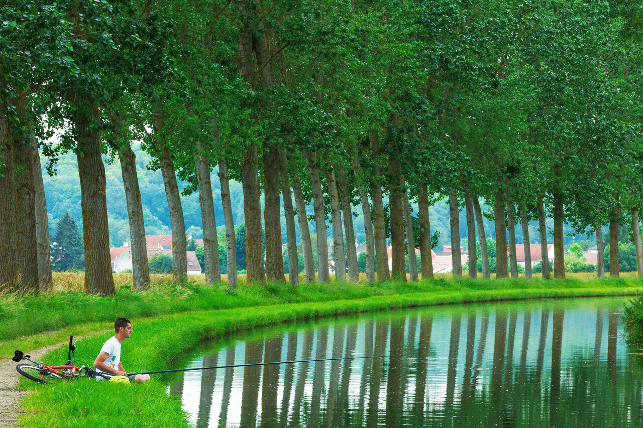 Man fishing by a canal with bicycle, green trees at CAPFUN Le Village Parisien campsite in VARREDDES (77).