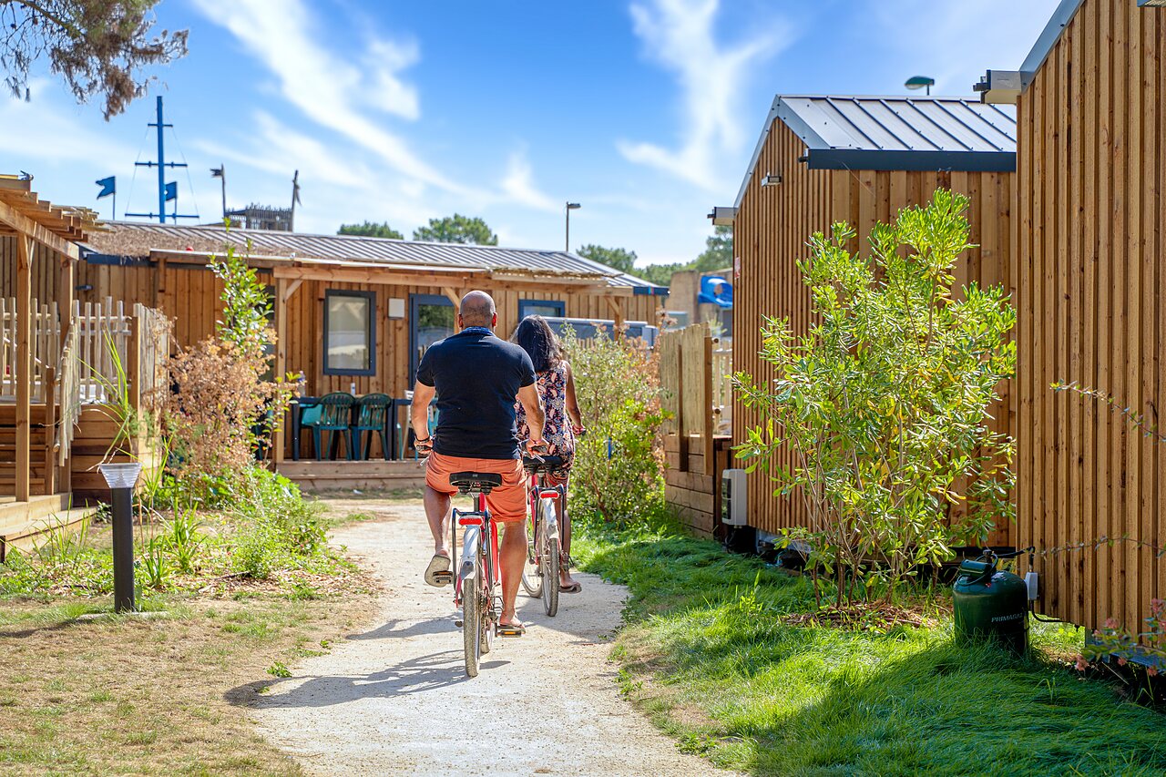 Couple cycling on a path between Mobile homes at CAPFUN Zagarella campsite in LONGEVILLE SUR MER (85).